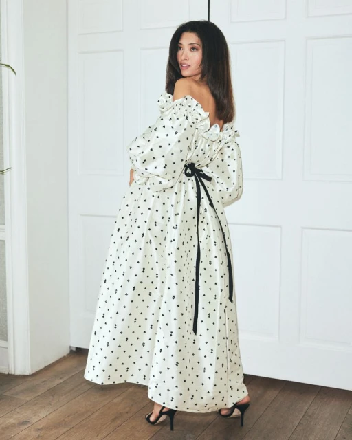 Elegant woman in off-shoulder dress with black ribbon detail poses indoors against a white paneled door and hardwood floor.