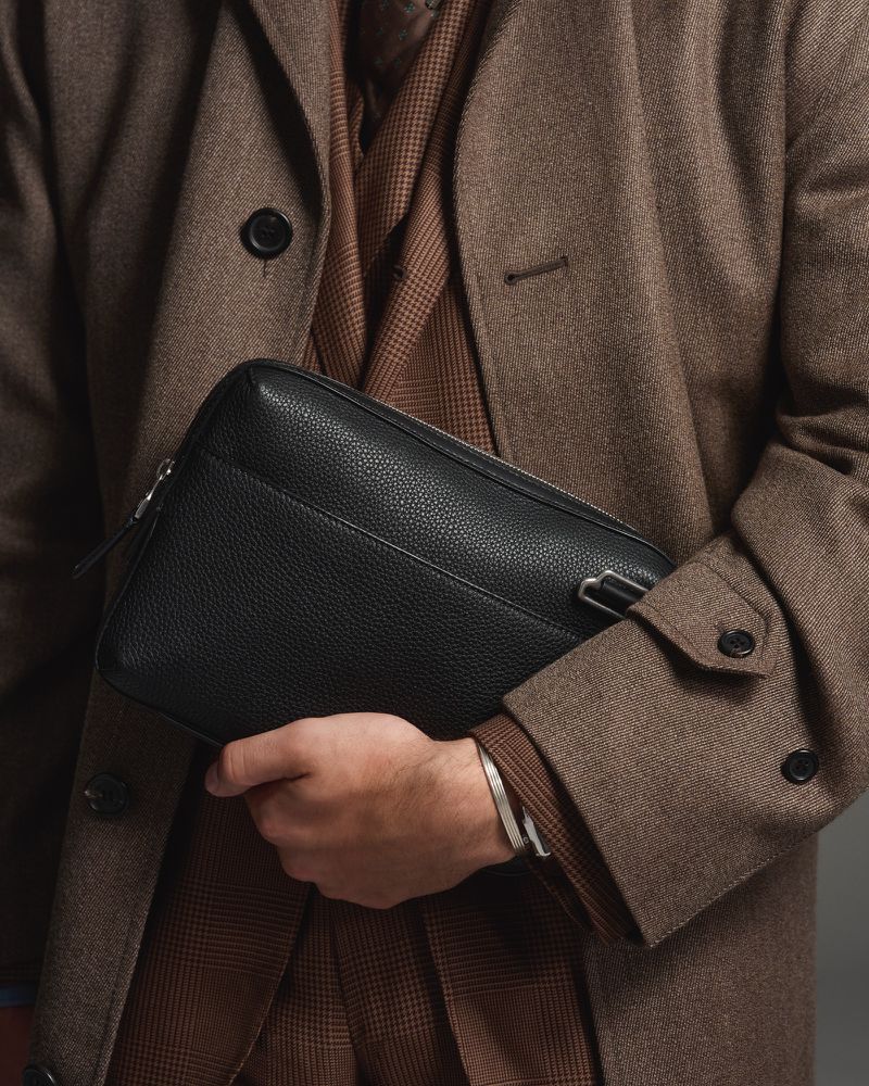 Close-up of adult male holding a black leather clutch bag, wearing a brown coat and a plaid suit, showcasing fashion and style.