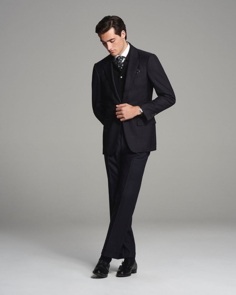 Man in a dark suit and patterned vest standing in a photo studio looking down. Classic formalwear for men.