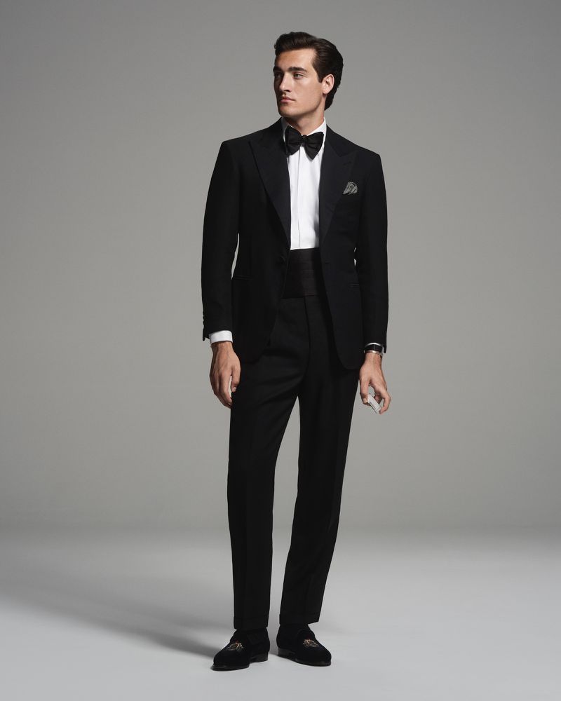 Man in black tuxedo with bow tie in a studio shot. He is wearing a white shirt and black dress pants with loafers. Formal fashion.