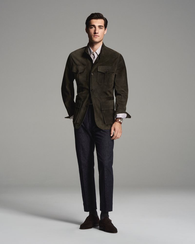 Full-length studio shot of an adult male model in a casual button-down shirt, olive green jacket, dark pants, and brown loafers.