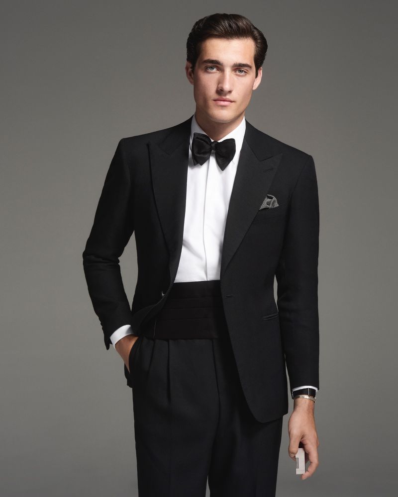 A handsome adult male in a black tuxedo and bow tie poses indoors against a grey backdrop, conveying elegance and formal style.