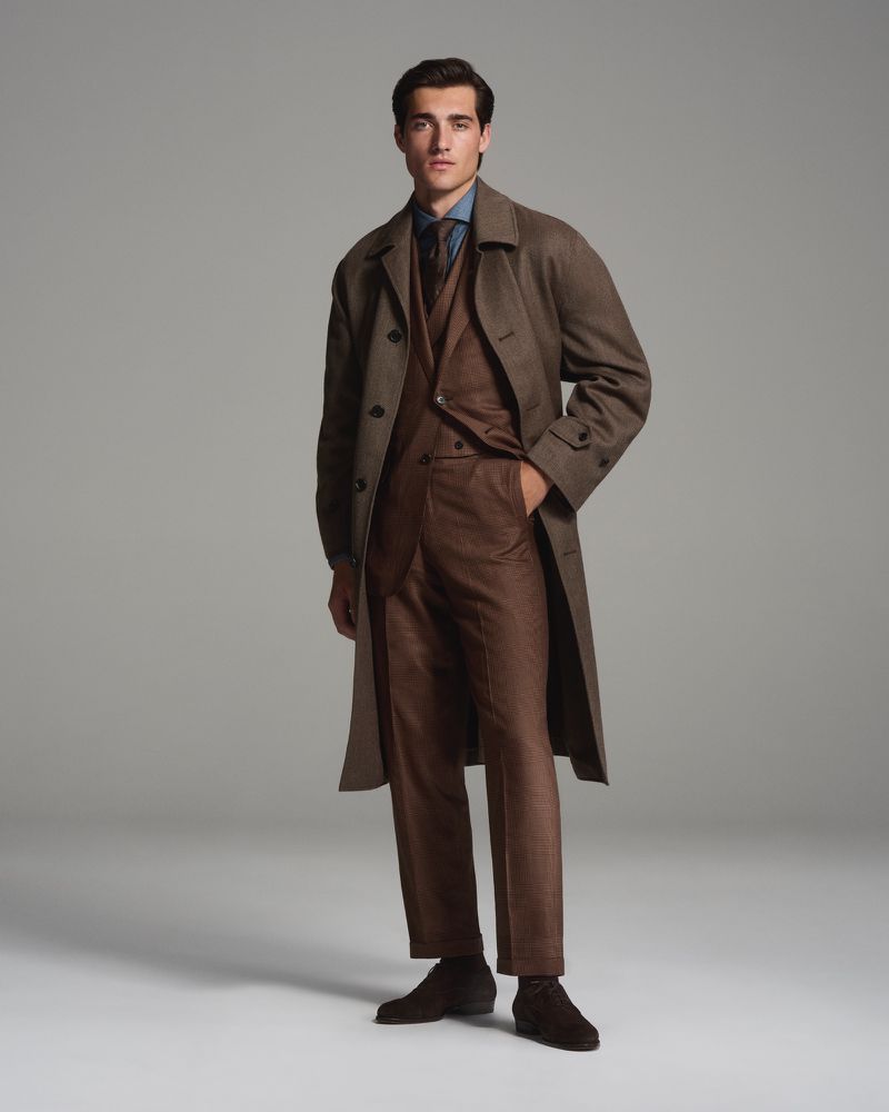 Full-length shot of a man in a three-piece suit with a long coat, posing in a studio with soft lighting on a neutral background.