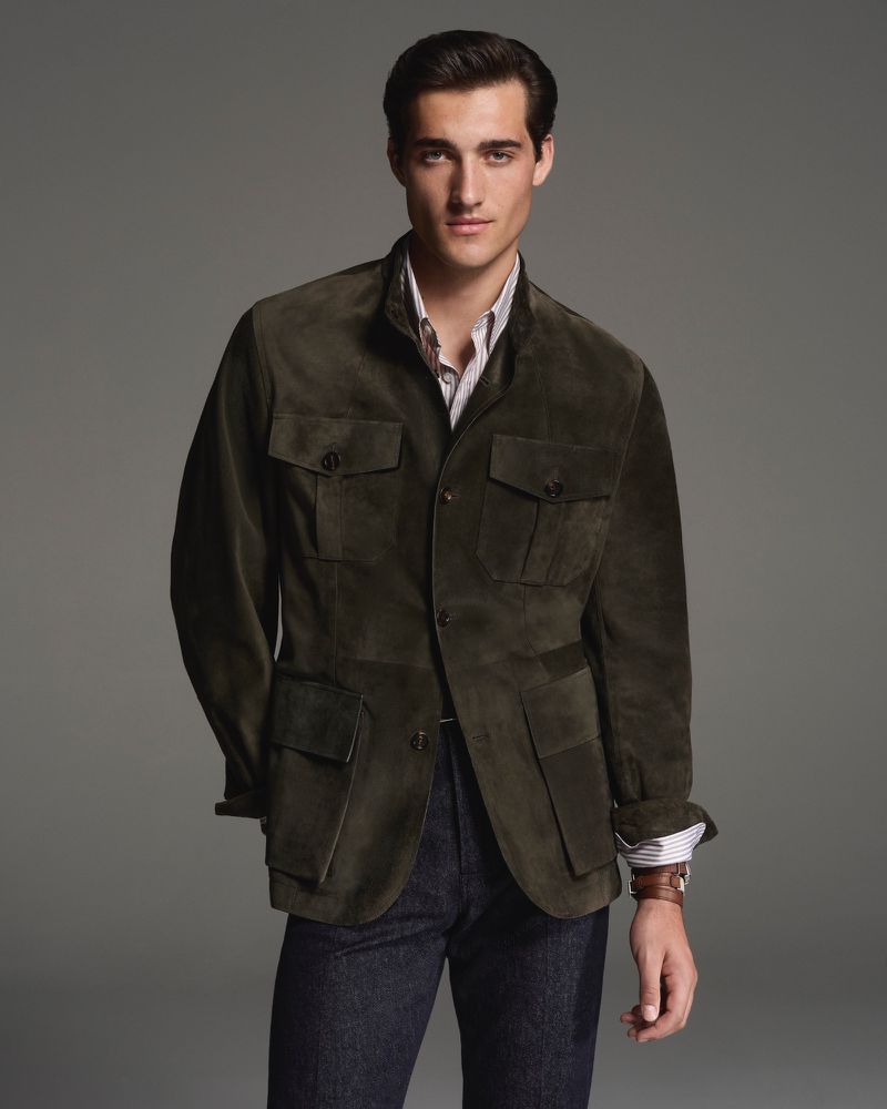 A man in a dark green suede jacket poses in a studio. His brown hair is neatly styled. He wears a striped shirt and dark jeans.