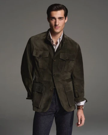 A man in a dark green suede jacket poses in a studio. His brown hair is neatly styled. He wears a striped shirt and dark jeans.