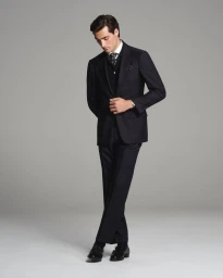 Model wearing a dark tailored suit and patterned tie standing against a plain grey studio background.