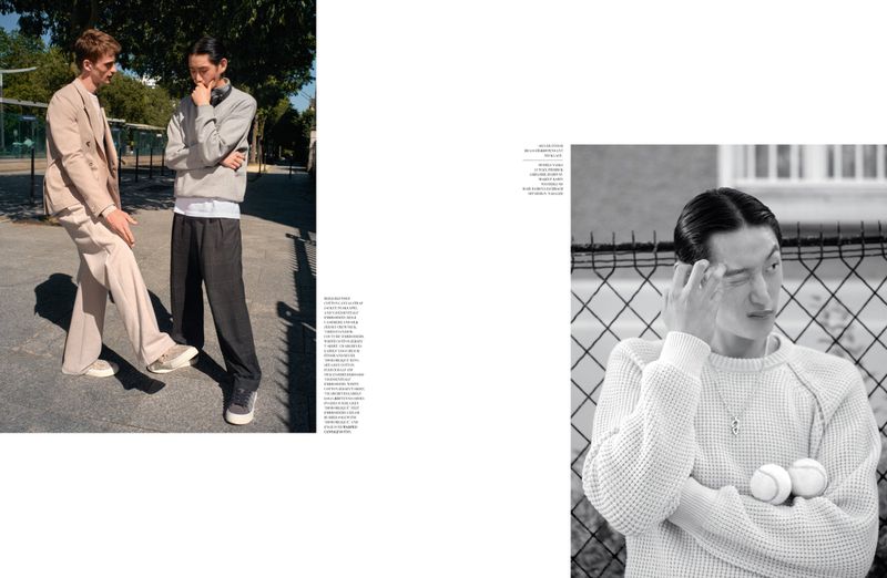 Two young adult men in stylish casual outfits pose on a paved surface with trees in the background in a fashion editorial style.