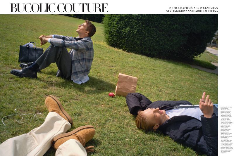 Two people relax on a grassy lawn. One man sits upright, while another lies down with a paper bag and an apple nearby.