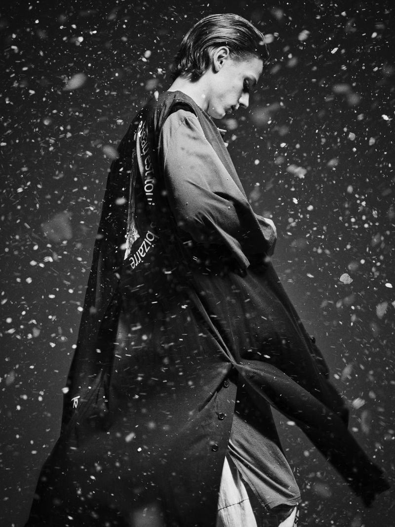 Black and white portrait of a slender adult male in a dark coat, looking down, with simulated snow falling against a dark background.