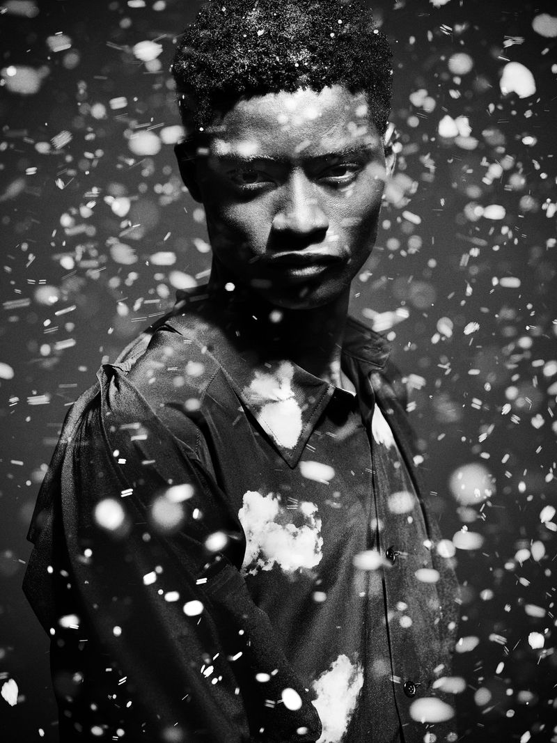 Black and white portrait of a young man with dark hair and a patterned shirt, surrounded by bokeh-like light particles.