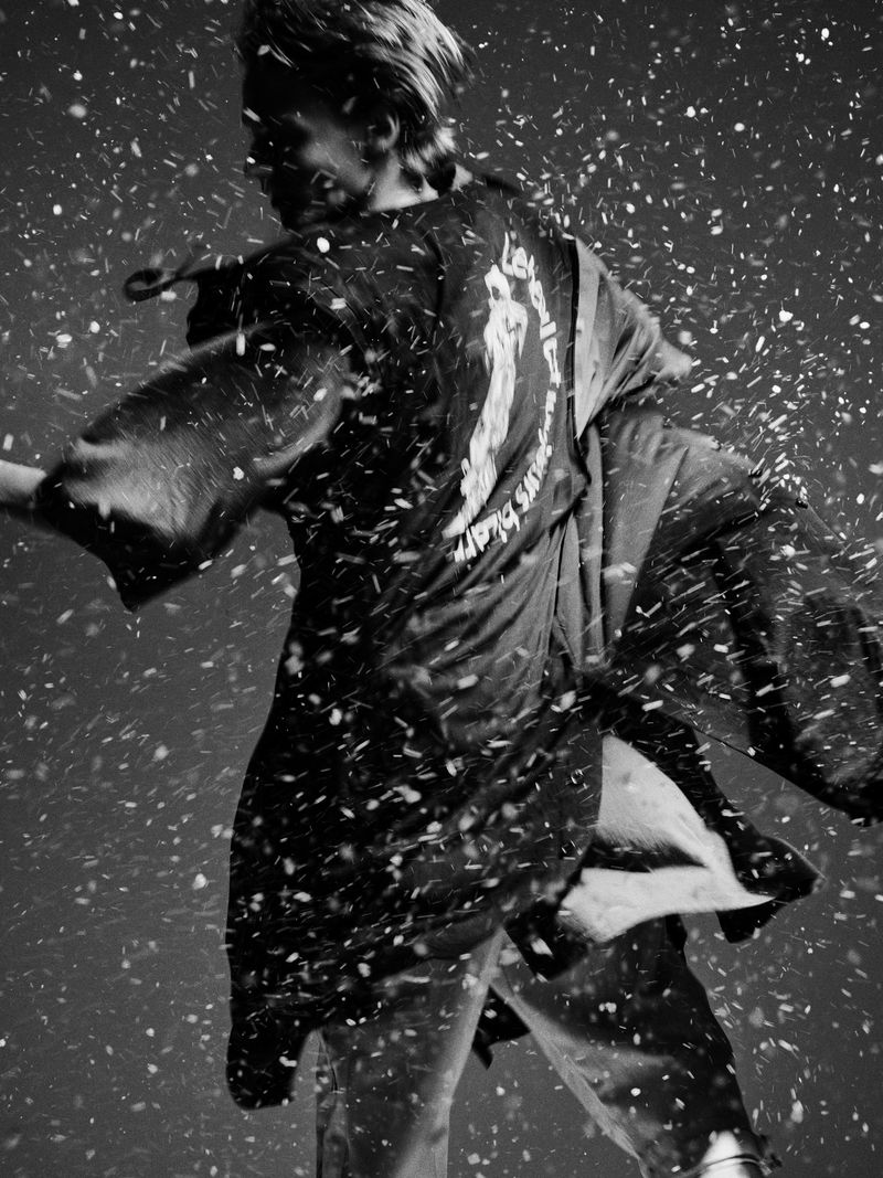 Monochrome photograph of an adult in a loose garment, possibly a robe, standing amid a flurry of light-colored particles, resembling snow or confetti.