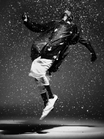 Black and white studio portrait of an adult wearing a jacket and athletic shorts, jumping in the air as if dancing in a snow storm.