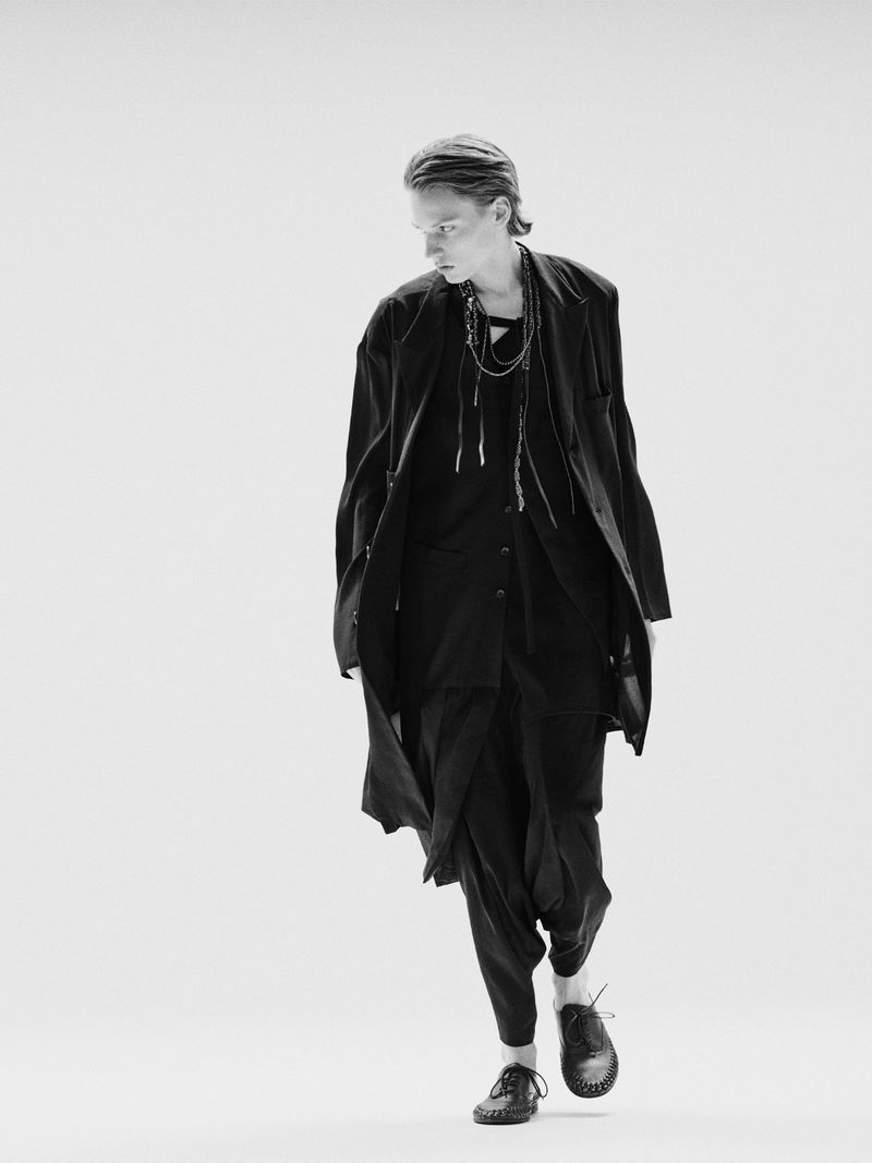 Monochrome fashion shot of a slender adult male wearing an open black long coat, shirt, and draped pants, walking on a white background.