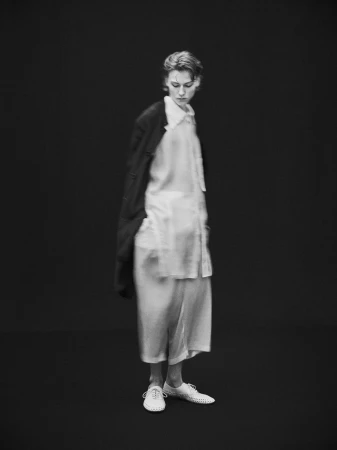 Black and white fashion shot of a model wearing a blouse, jacket, and slacks against a dark backdrop in a photo studio.