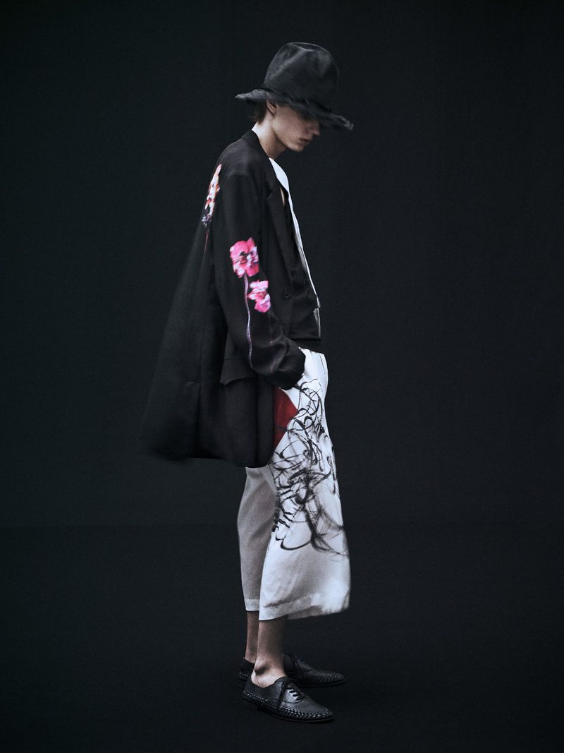 Fashionable adult male in dark hat, black coat with floral design, and flowing pants stands against a dark studio backdrop, side view.