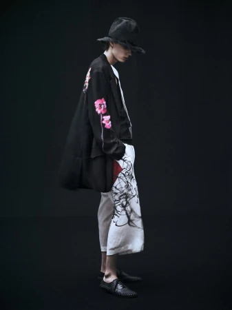 Fashionable adult male in dark hat, black coat with floral design, and flowing pants stands against a dark studio backdrop, side view.