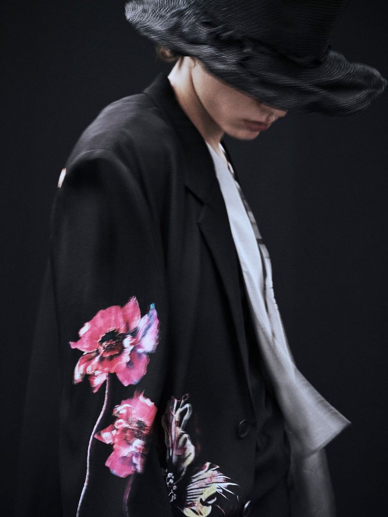 Fashion shot of an adult wearing a black blazer with floral design, a black hat, and a gradient shirt against a dark backdrop.