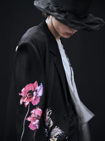 Fashion shot of an adult wearing a black blazer with floral design, a black hat, and a gradient shirt against a dark backdrop.