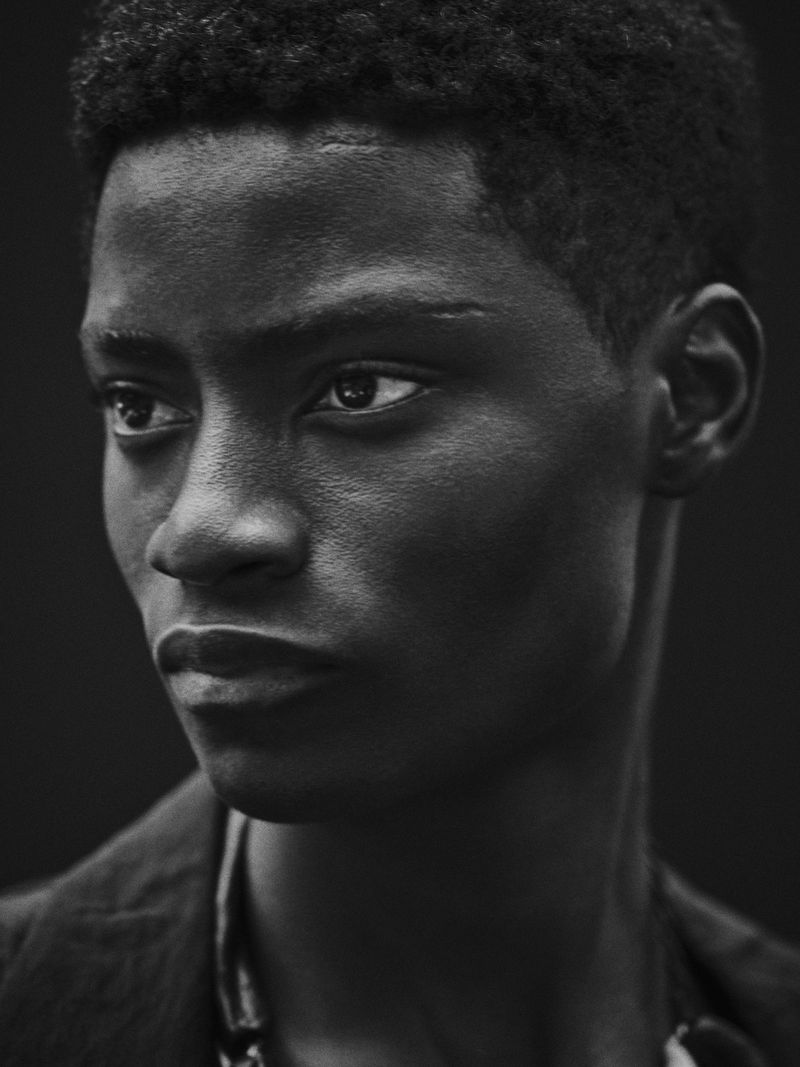 Monochromatic portrait of a young Black man with short hair, wearing a dark jacket and light shirt against a dark backdrop.