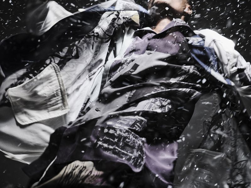 Low-angle shot of a male model in layered patterned clothing amidst falling white particles against a dark background