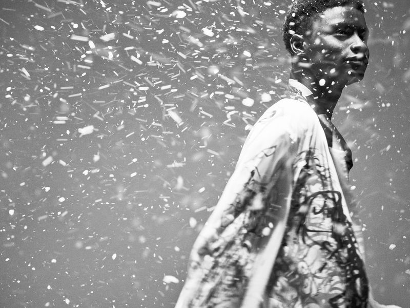 Portrait of adult male in patterned jacket with falling snow, captured in black and white with shallow depth of field in outdoor setting.