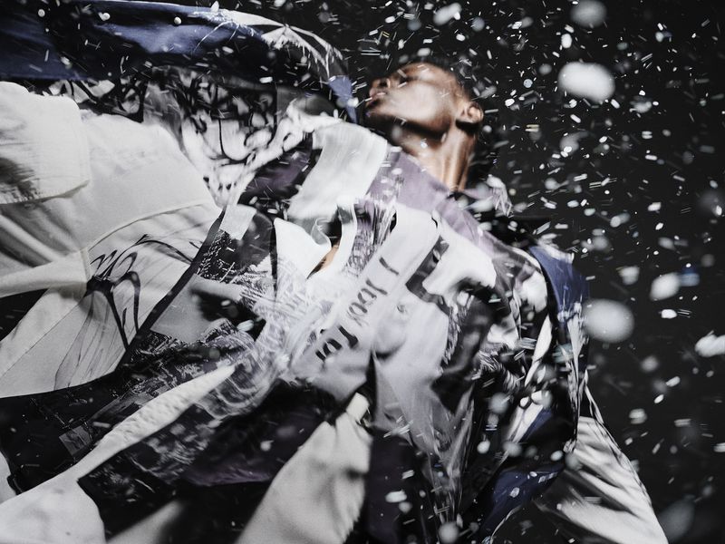 Fashion shot of an adult with light shining on their face, wearing a jacket with graphic prints, while snow particles surround them.