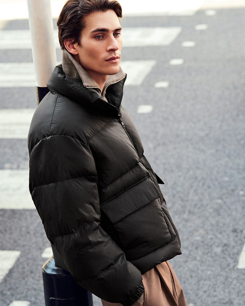 Male model in a puffer jacket leans against a pole in an urban setting, showcasing fashion and outdoor lifestyle photography.