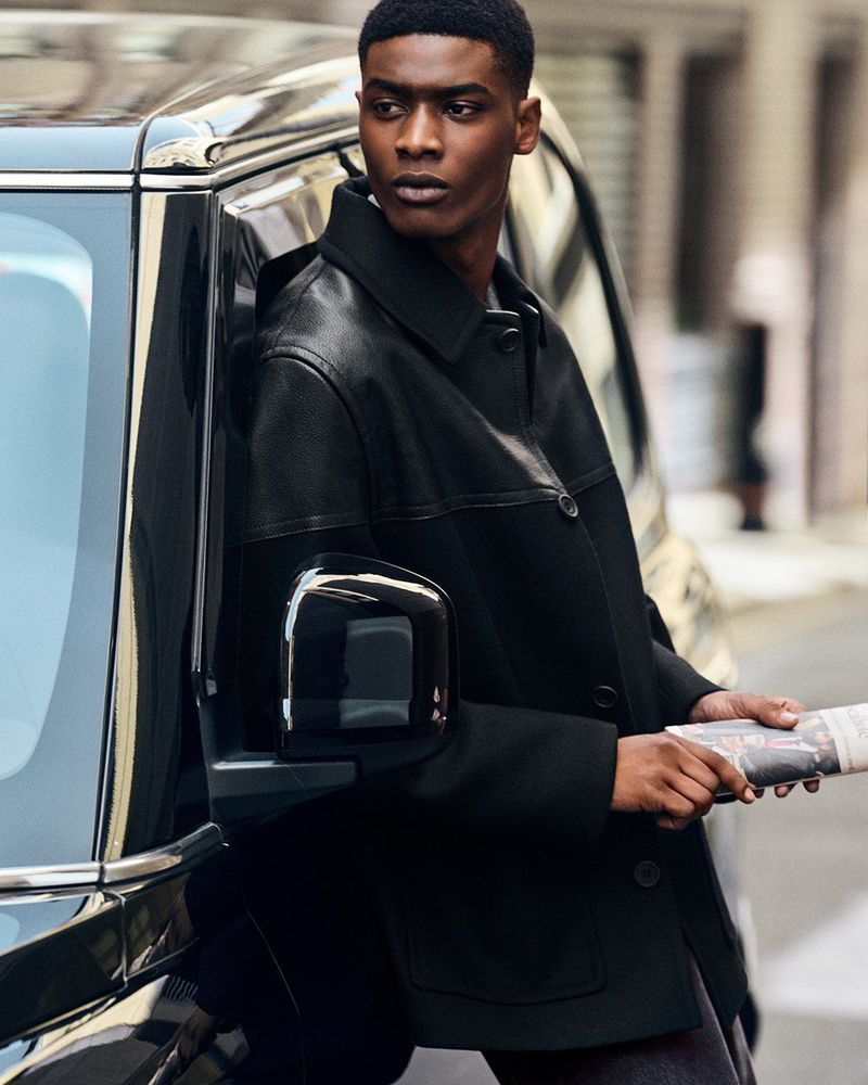 A Black male model leans against a black SUV on a city street, holding a newspaper, wearing a black leather and wool jacket, looking away.
