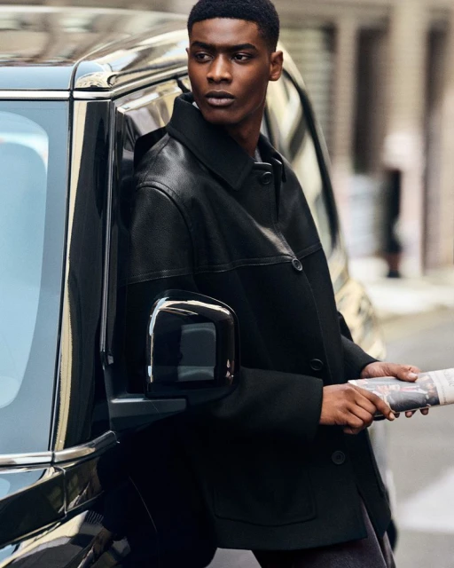 A Black male model leans against a black SUV on a city street, holding a newspaper, wearing a black leather and wool jacket, looking away.