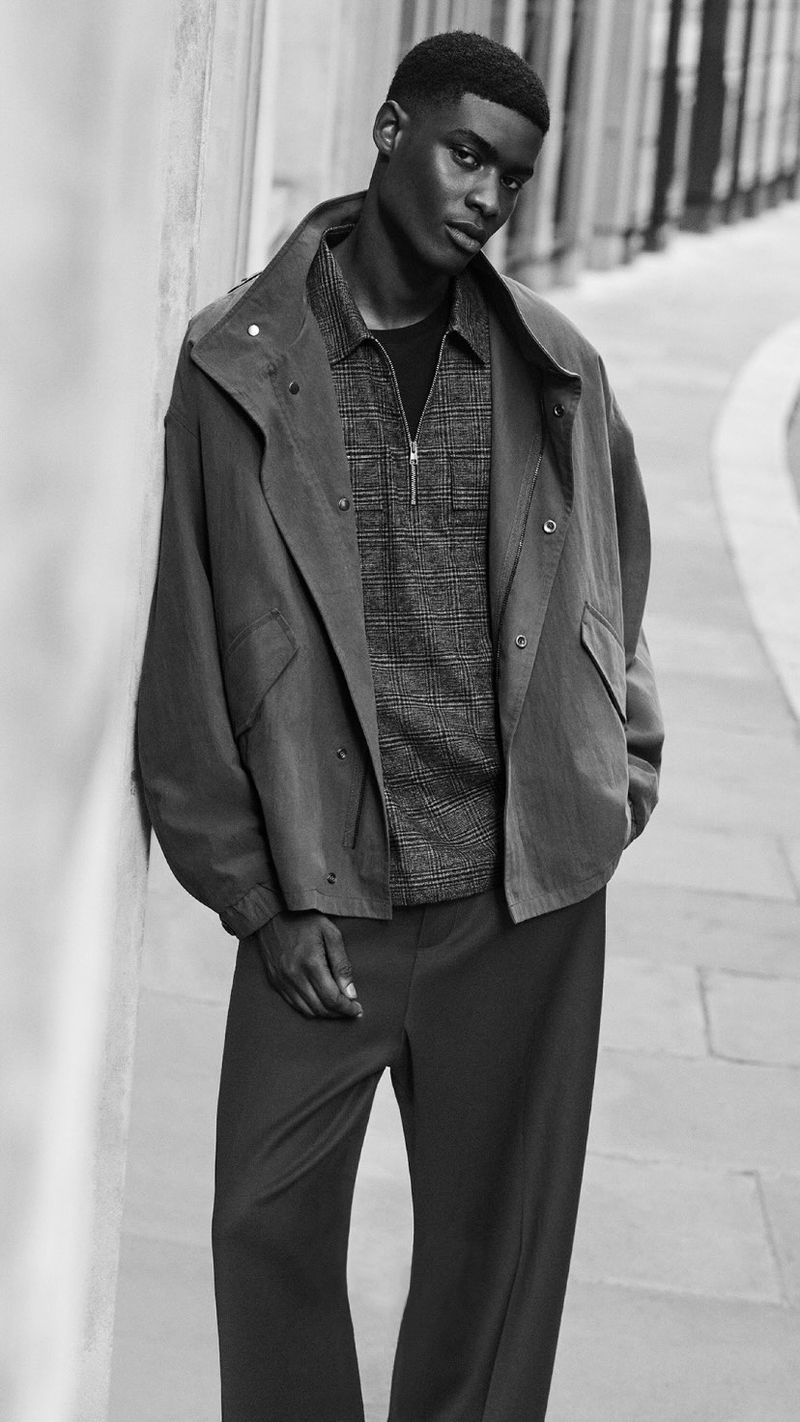 Black and white portrait of a male model leaning against a textured wall wearing a structured jacket and patterned zip shirt for COS