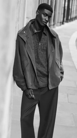 Black and white portrait of a male model leaning against a textured wall wearing a structured jacket and patterned zip shirt for COS