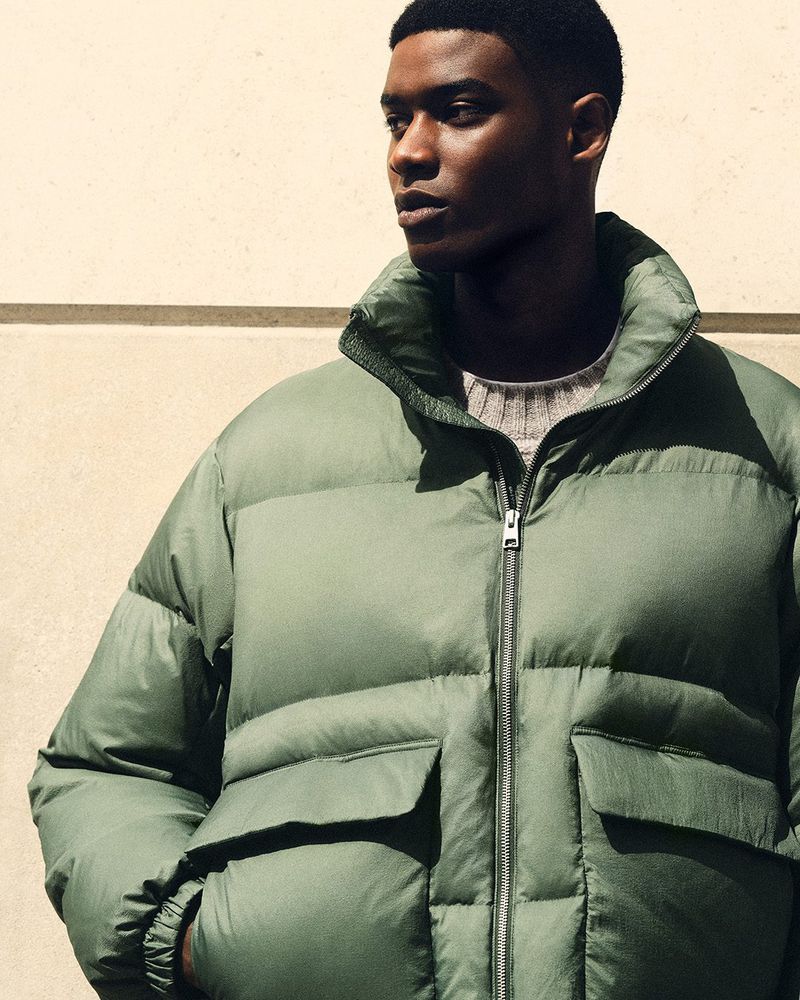 Close-up studio shot of a Black male model in an olive green puffer jacket and grey crew neck sweater against a neutral wall.