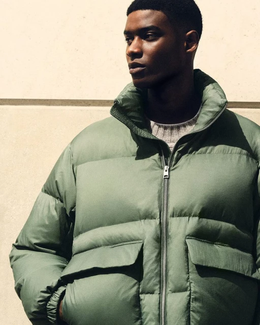 Close-up studio shot of a Black male model in an olive green puffer jacket and grey crew neck sweater against a neutral wall.