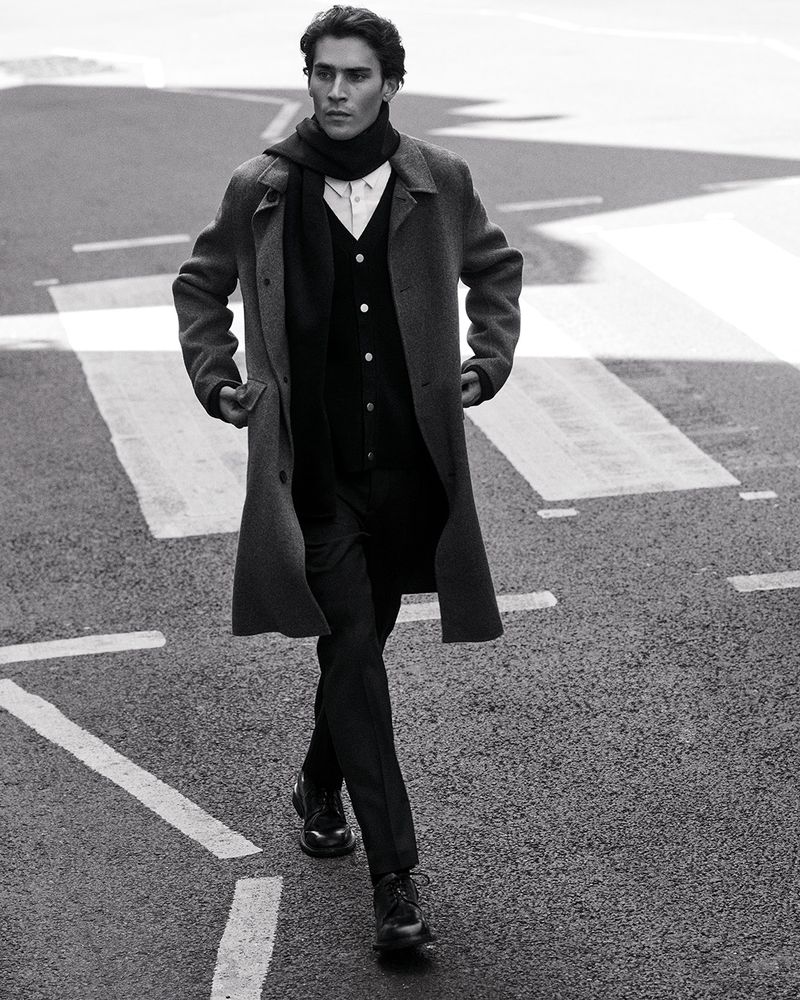 Man wearing a grey wool coat and scarf walking on an asphalt street with road markings.