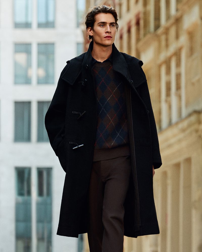 Male model wearing a black duffle coat over an argyle sweater walking along a city street for COS menswear