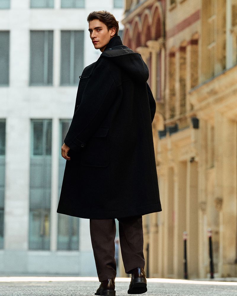 A man with dark hair, viewed from the back, walks along a city street wearing a black duffel coat, brown pants, and black boots.