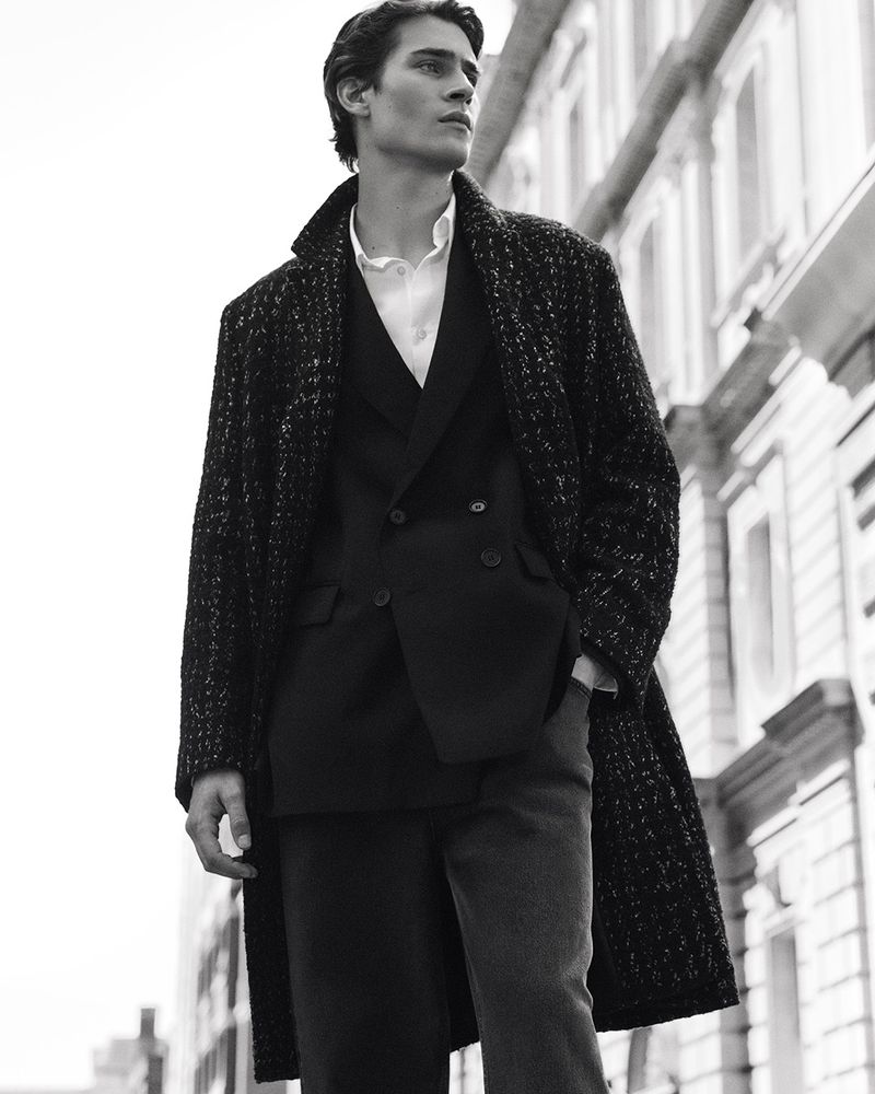 Black-and-white outdoor portrait of a tall, slender man wearing a textured overcoat and suit, standing in front of a building.