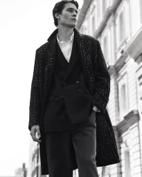 Monochrome fashion portrait of a male model wearing a dark textured coat and blazer standing outdoors