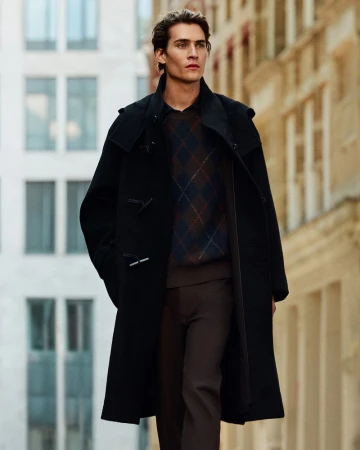 Male model wearing a black duffle coat over an argyle sweater walking along a city street for COS menswear