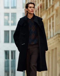 Male model wearing a black duffle coat over an argyle sweater walking along a city street for COS menswear