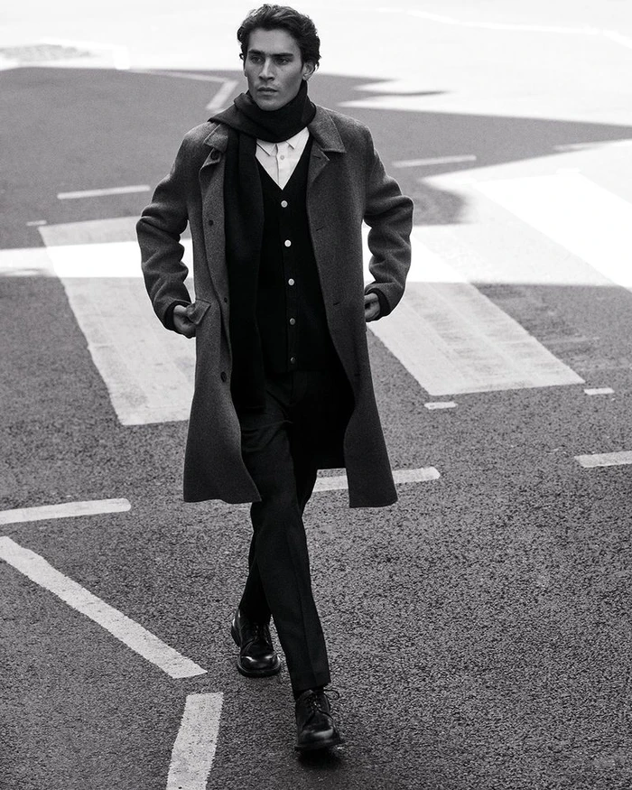 Man wearing a grey wool coat and scarf walking on an asphalt street with road markings.