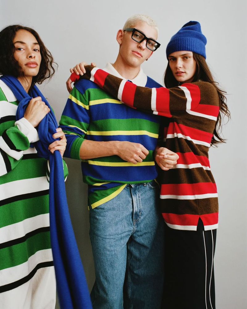 Three fashion models in striped shirts and blue accessories pose in a studio with soft lighting. One wears glasses and denim jeans.
