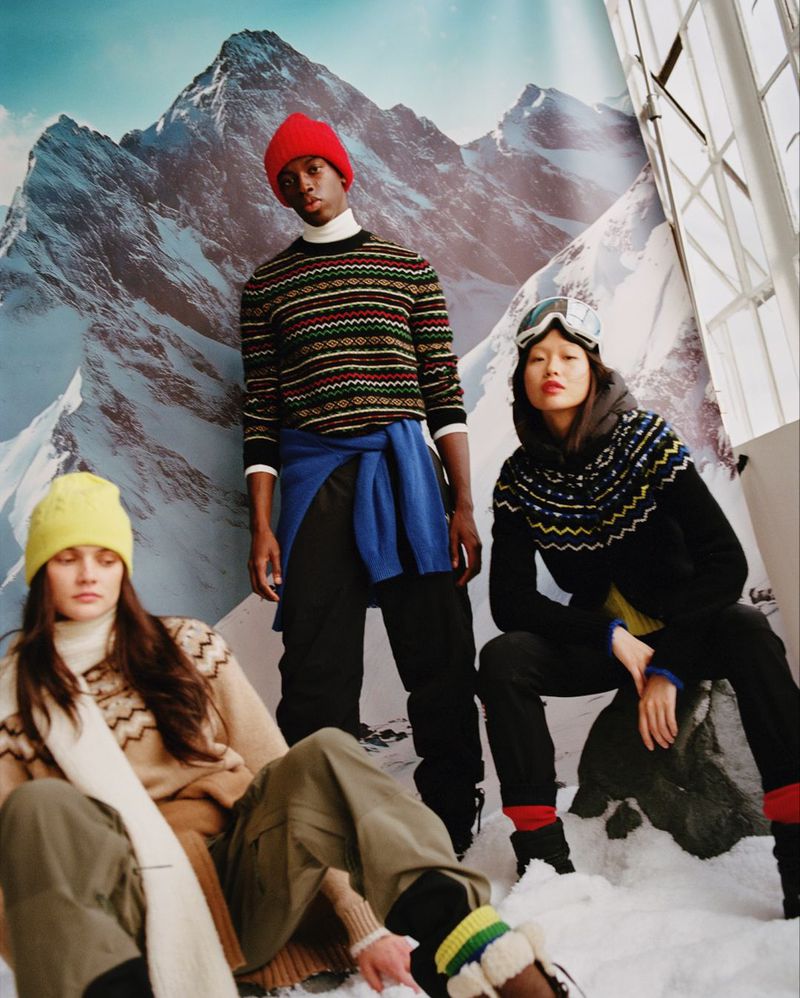 Group of three fashion models posing in winter apparel with a mountain backdrop, showcasing knitted sweaters, hats, and goggles.