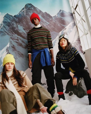 Group of three fashion models posing in winter apparel with a mountain backdrop, showcasing knitted sweaters, hats, and goggles.