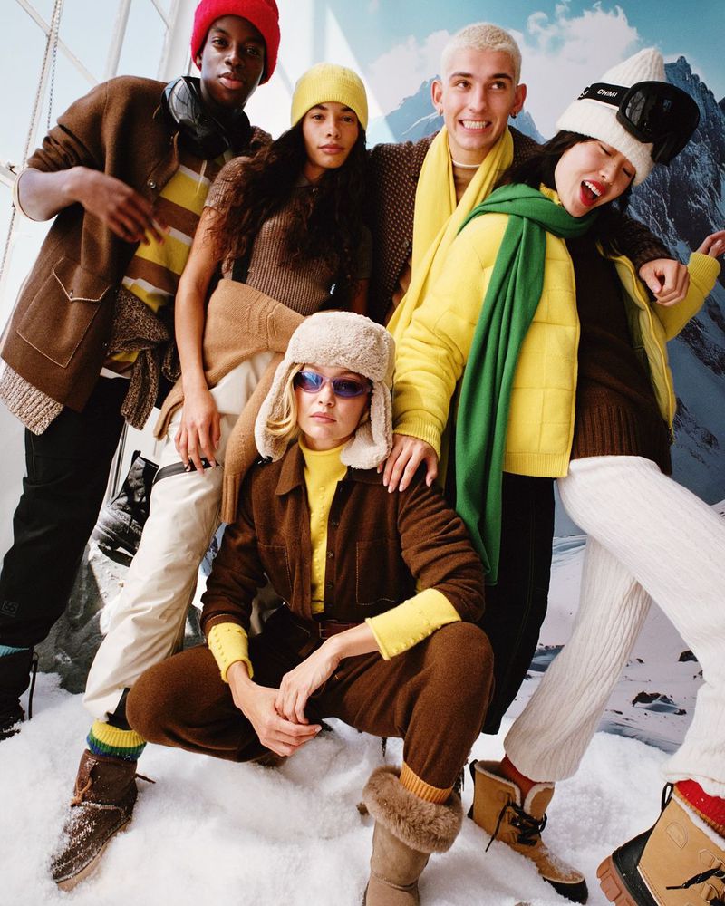Group of five young adult fashion models in winter clothing posing in an indoor studio with a backdrop of snow-capped mountains.