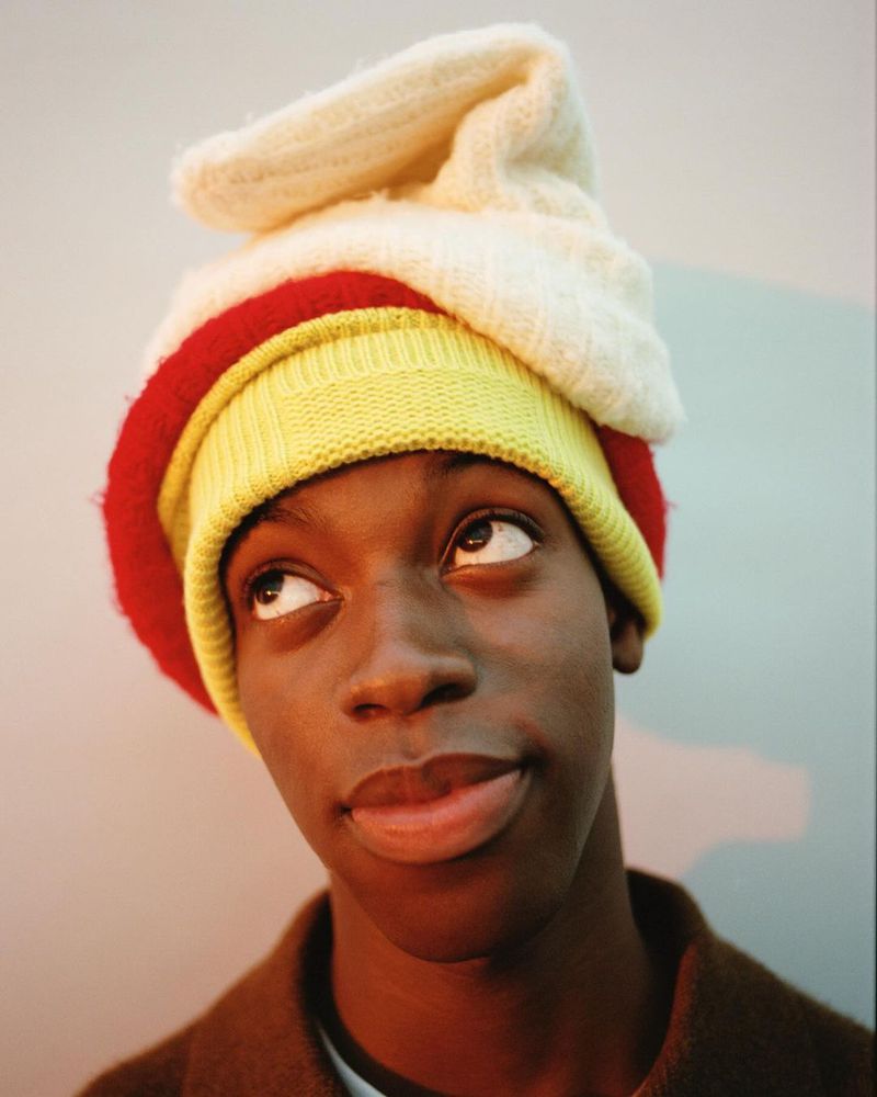 Person with an upward gaze wearing a layered hat in yellow, red, and white, with a brown jacket against a neutral gradient background.