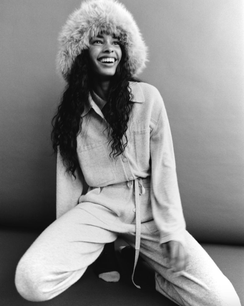 A smiling woman with dark hair, wearing a fur hat and a casual suit, looks off-camera in a monochrome studio portrait.