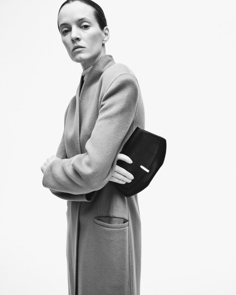 Model Daria Strokous holding a black handbag while wearing a long coat against a white background