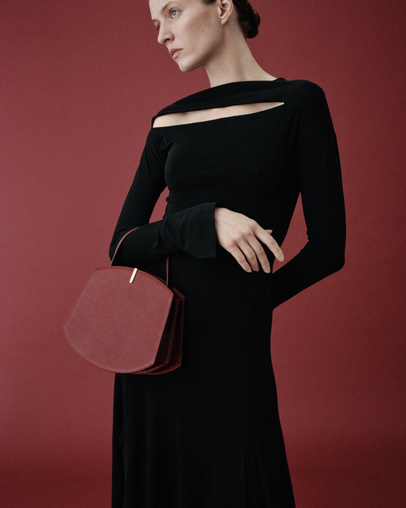Elegant woman poses in black long-sleeve dress against a red background, holding a unique geometric purse. Fashion editorial aesthetic.