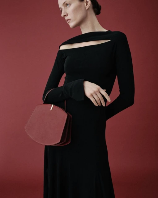 Elegant woman poses in black long-sleeve dress against a red background, holding a unique geometric purse. Fashion editorial aesthetic.